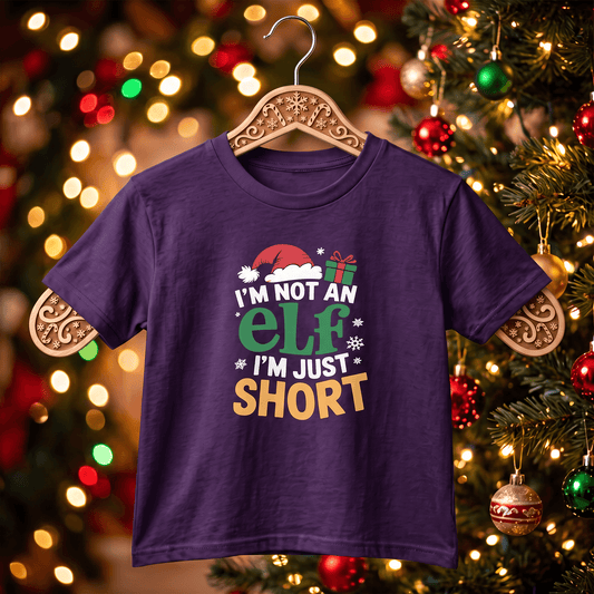 A dark purple kids' t-shirt with a Christmas graphic reading 'I'M NOT AN ELF I'M JUST SHORT' in green, yellow, and white, hanging on a wooden hanger against a blurred Christmas tree background.