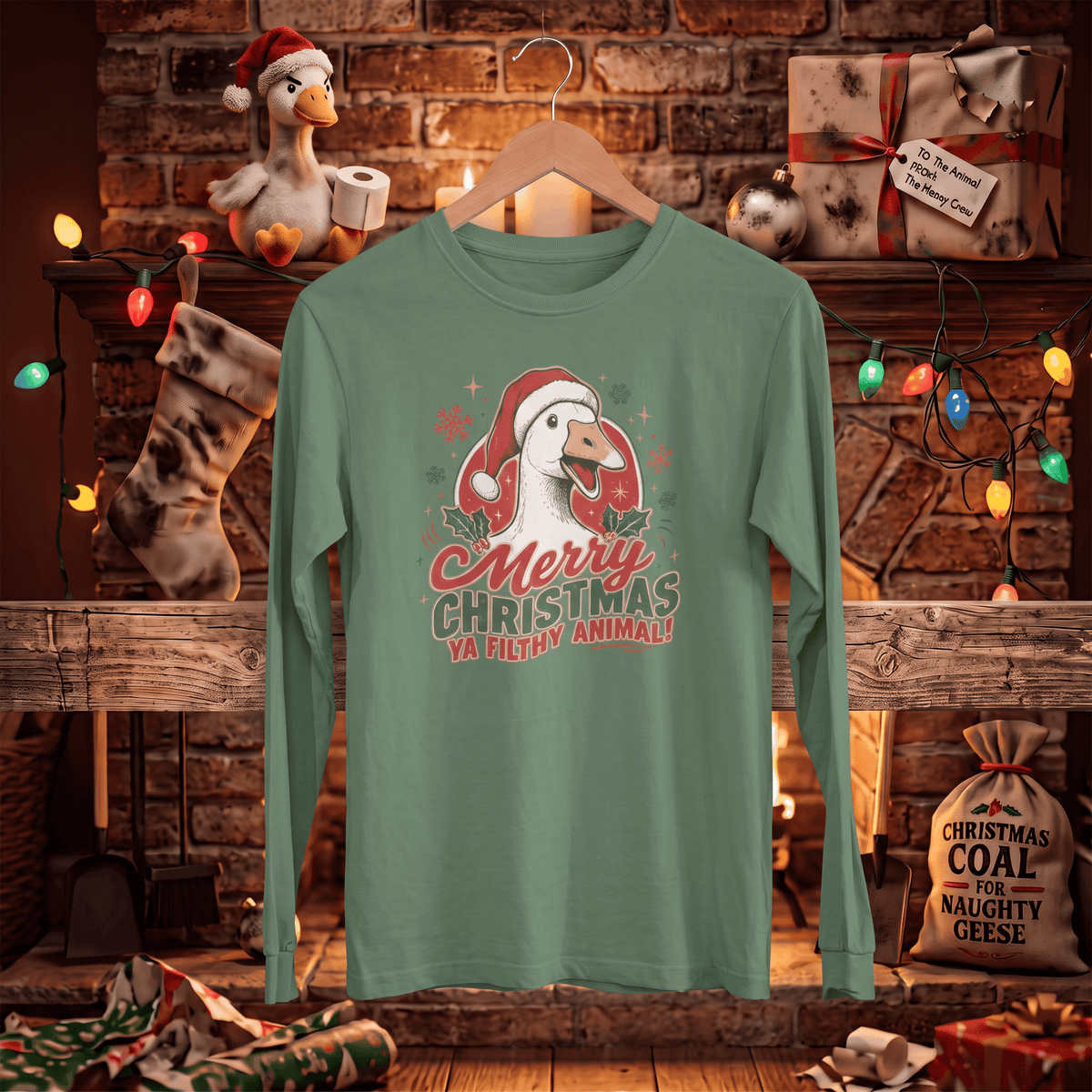 A sage green long-sleeve t-shirt with a 'Merry Christmas Ya filthy animal!' goose graphic, hanging on a wooden hanger in front of a cozy, warmly lit Christmas fireplace background.