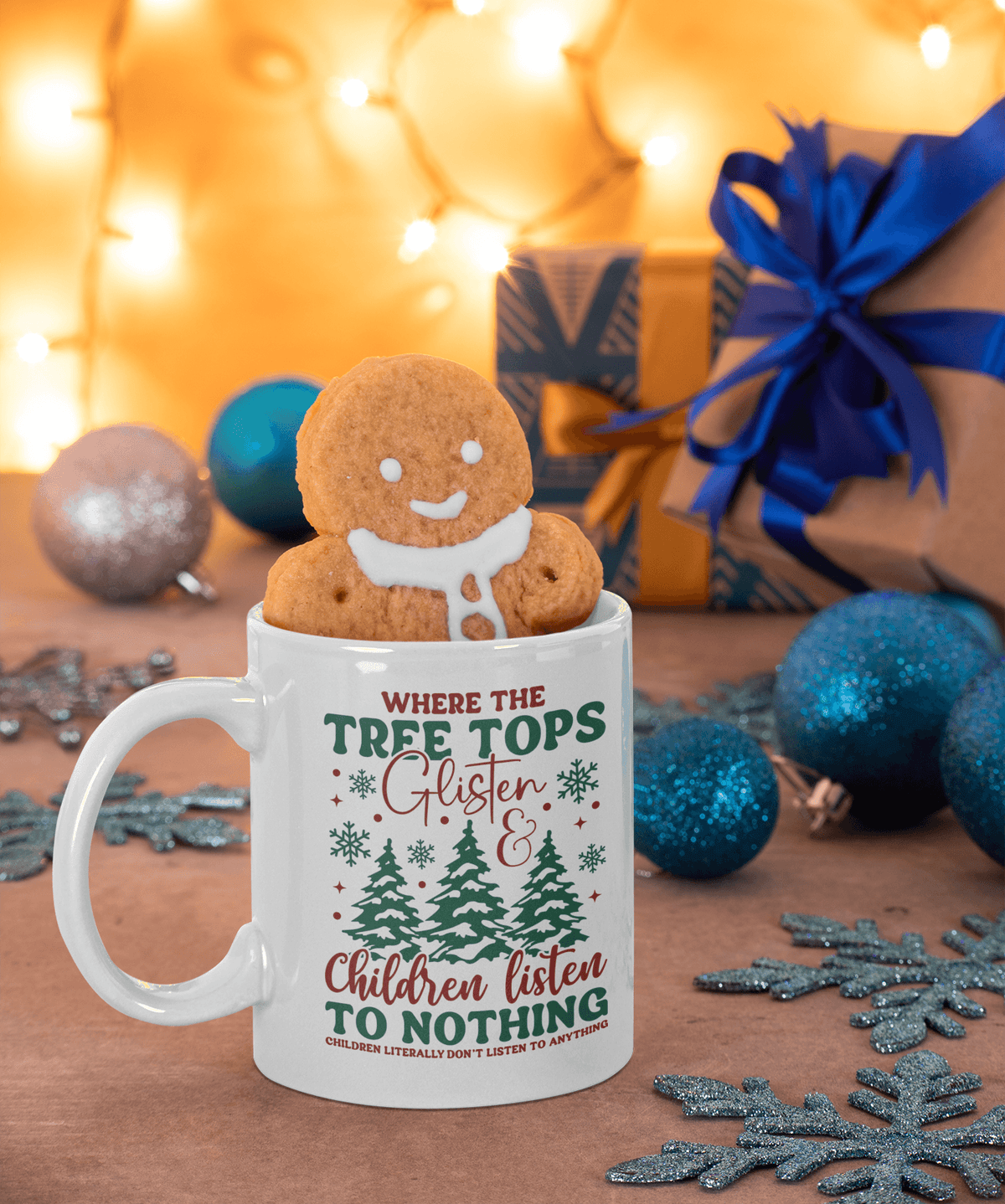 A white ceramic mug with a gingerbread cookie peeking out from the top, styled on a wooden surface with Christmas ornaments, gift boxes, and warm string lights in the background. The mug features a festive holiday graphic with the text 'WHERE THE TREE TOPS Glister & Children listen to NOTHING'.