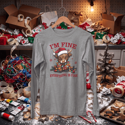 A light gray long-sleeve t-shirt with a Christmas Highland cow graphic and the text 'I'M FINE / EVERYTHING IS FINE' hanging on a wooden hanger. The shirt is displayed in a messy, cluttered holiday workshop environment with wrapping paper, boxes, and lights.