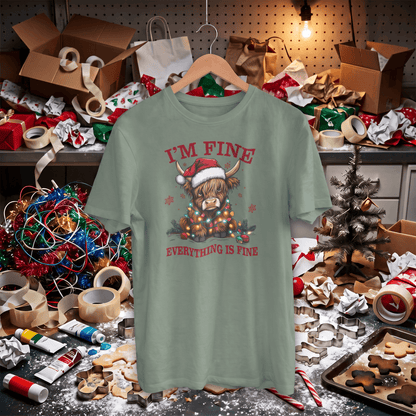 A sage green graphic t-shirt on a wooden hanger, featuring a highland cow wearing a Santa hat and scarf with the text 'I'M FINE' and 'EVERYTHING IS FINE' in red. The shirt is displayed in a messy, cluttered Christmas workshop setting with wrapping paper, boxes, and holiday decorations.