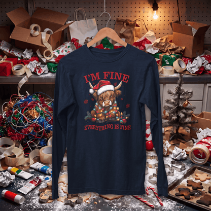 A navy blue long-sleeve t-shirt with a festive graphic of a Highland cow under Christmas lights and the text 'I'M FINE / EVERYTHING IS FINE' in red, hanging on a wooden hanger. The shirt is displayed in a cluttered, festive workshop or bakery environment with wrapping paper, lights, and cookies.