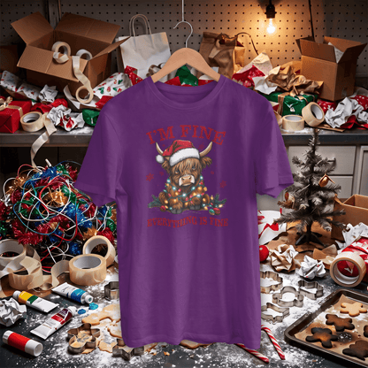 A purple short-sleeve graphic t-shirt on a wooden hanger, featuring a Highland cow in a Santa hat surrounded by Christmas lights and the text 'I'M FINE' and 'PERFECTLY FINE IS NOT'. The shirt is displayed in a cluttered, festive craft workspace filled with boxes, tape, holiday lights, and cookie cutters.