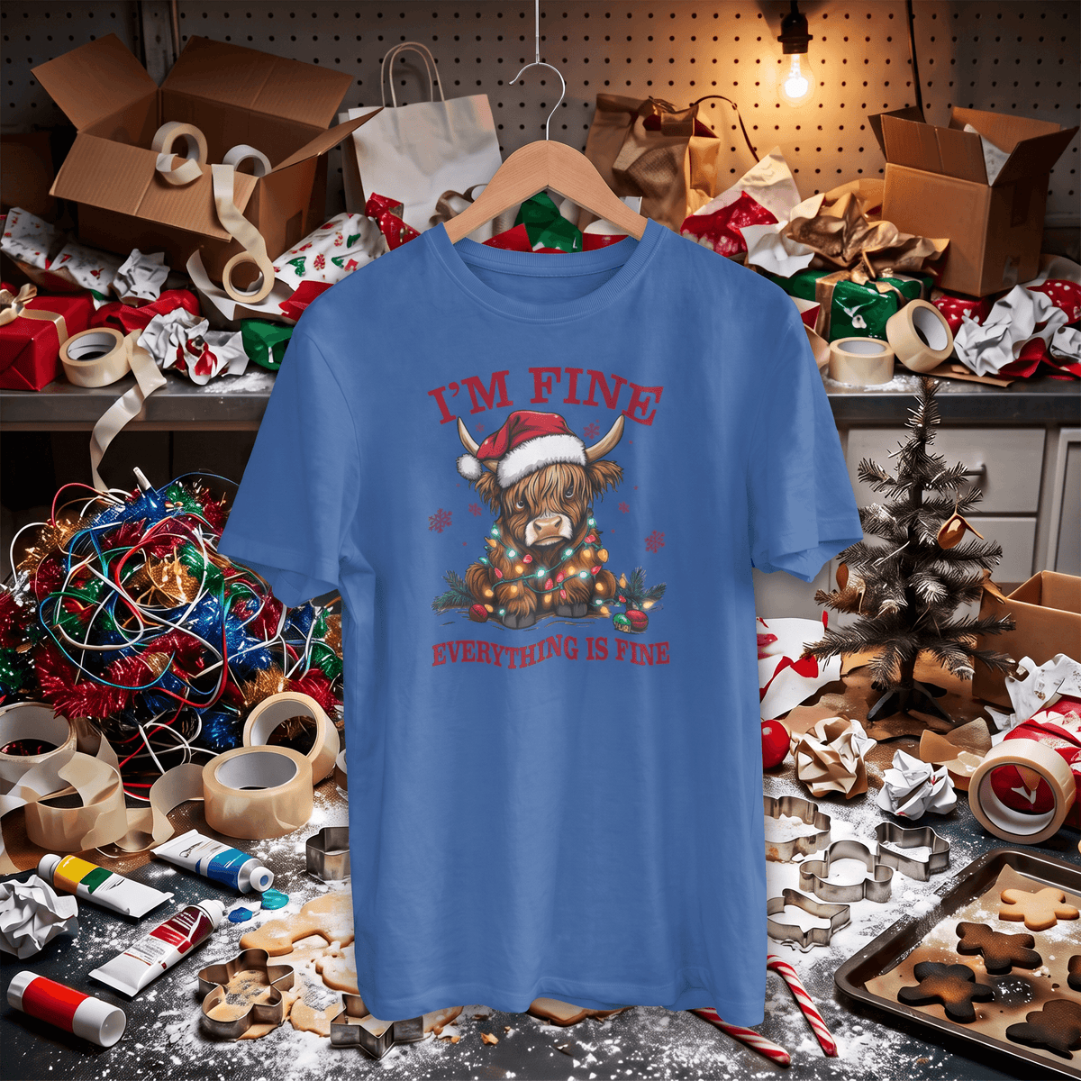 A blue graphic t-shirt hanging on a wooden hanger in a cluttered, festive Christmas workshop setting. The shirt features a highland cow wearing a Santa hat and scarf, surrounded by ornaments, with the text 'I'M FINE' and 'EVERYTHING IS FINE' in red.