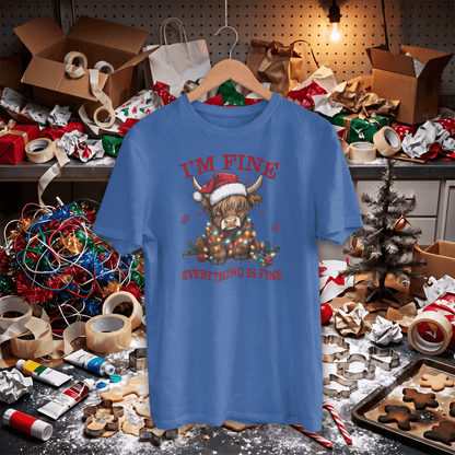 A blue graphic t-shirt hanging on a wooden hanger in a cluttered, festive Christmas workshop setting. The shirt features a highland cow wearing a Santa hat and scarf, surrounded by ornaments, with the text 'I'M FINE' and 'EVERYTHING IS FINE' in red.