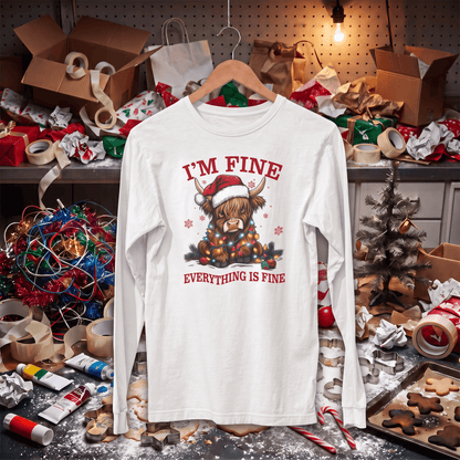 A white long-sleeve t-shirt with a Christmas Highland cow graphic and the text 'I'M FINE / EVERYTHING IS FINE' hanging on a wooden hanger. The shirt is displayed in a messy, cluttered holiday workshop environment with wrapping paper, lights, and craft supplies.