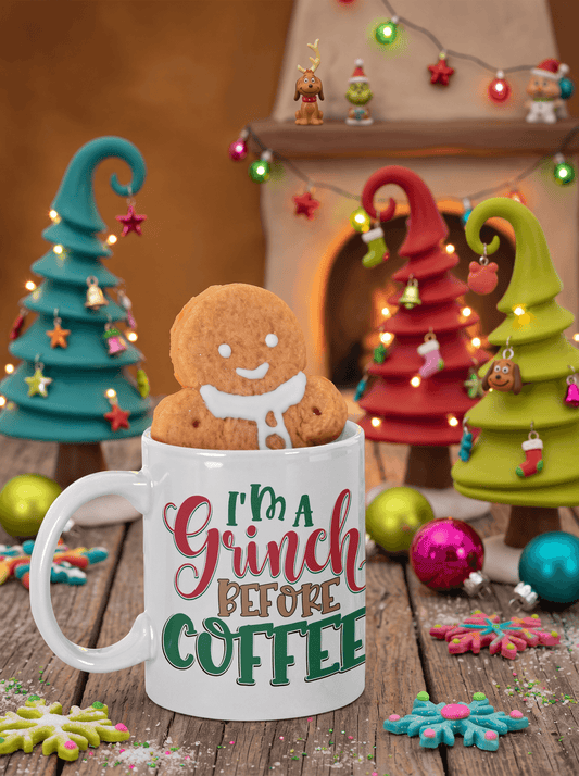 A white ceramic mug with the text 'I'm A Grinch BEFORE COFFEE' sits on a rustic wooden table, with a gingerbread man cookie peeking out from above the mug. The background is a cozy holiday scene with miniature Christmas trees, warm lights, ornaments, and a fireplace.