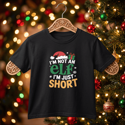 A black short-sleeve t-shirt hanging on a decorative wooden hanger, featuring a Christmas graphic that reads 'I'M NOT AN ELF I'M JUST SHORT' in white, green, and gold lettering. The background shows a blurred Christmas tree with warm lights and ornaments.