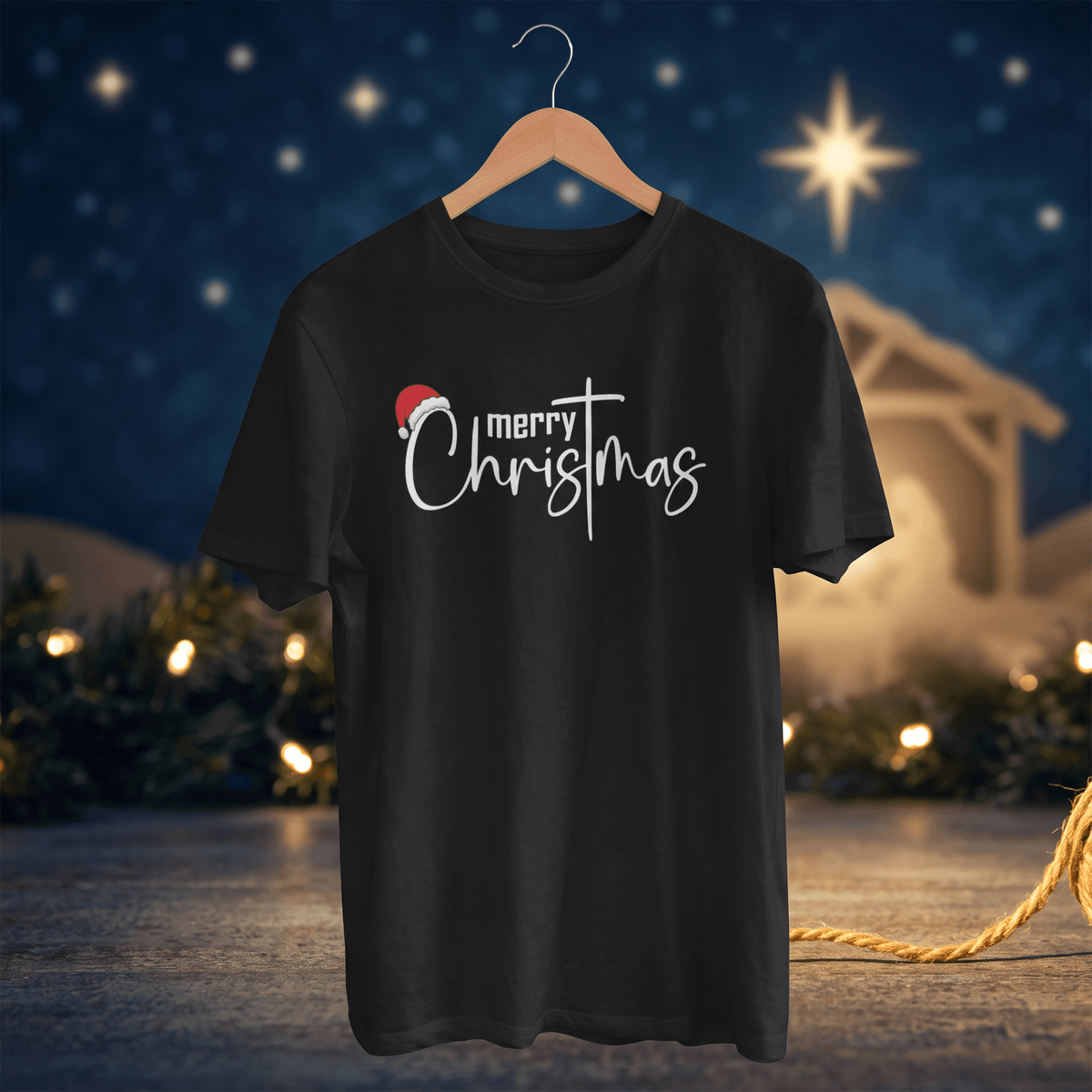 A black short-sleeve t-shirt on a wooden hanger, featuring a 'merry Christmas' graphic with a small red Santa hat, set against a blurred Christmas manger night scene background.