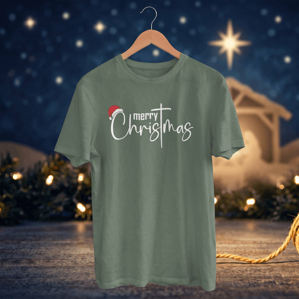 A sage green short-sleeve t-shirt on a wooden hanger, featuring a 'merry Christmas' graphic with a small red Santa hat, set against a festive Christmas night background with lights and a star.