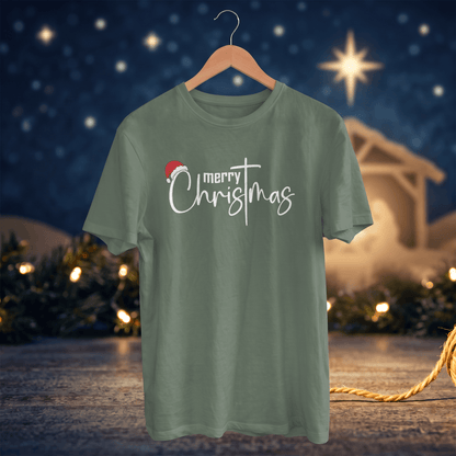A sage green short-sleeve t-shirt on a wooden hanger, featuring a 'merry Christmas' graphic with a small red Santa hat, set against a festive Christmas night background with lights and a star.
