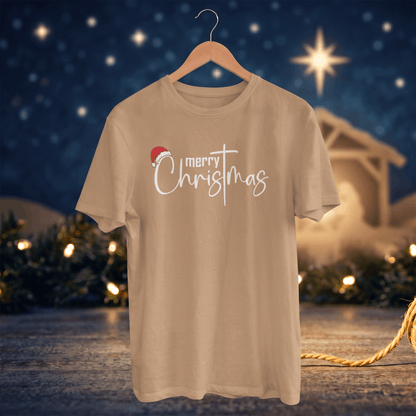 A beige short-sleeve t-shirt on a wooden hanger, featuring a 'merry Christmas' graphic with a small red Santa hat. The background is a festive, blurred Christmas scene with a starry night sky, lights, and a stable.