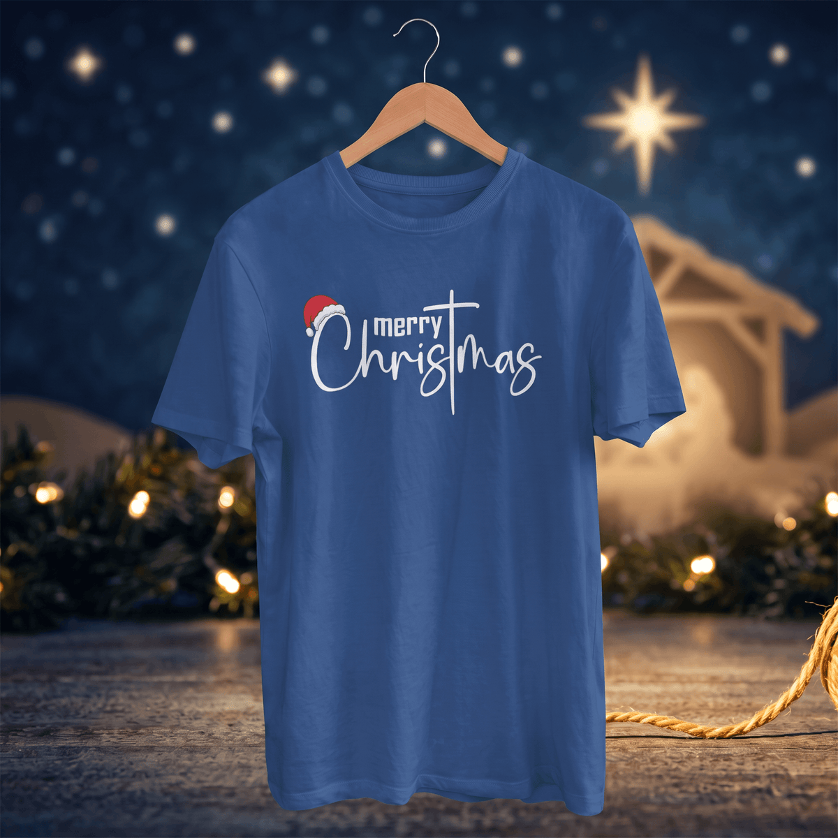 A royal blue short-sleeve t-shirt on a wooden hanger, featuring a 'merry Christmas' graphic with a small red Santa hat, set against a festive Christmas night background with lights and a star.