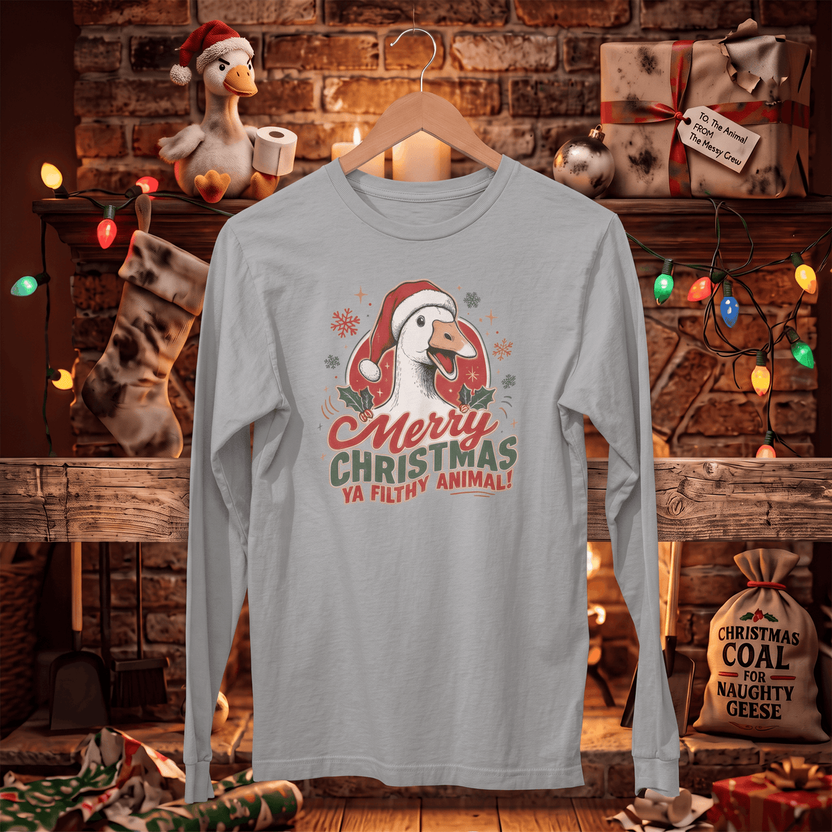 A light gray long-sleeve t-shirt with a Christmas goose graphic and the text 'Merry Christmas Ya filthy animal!' hanging on a wooden hanger in front of a cozy, decorated fireplace scene.