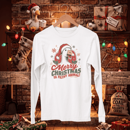 A white long-sleeve t-shirt with a Christmas goose graphic hanging on a wooden hanger in front of a cozy, decorated fireplace. The graphic features a cartoon goose wearing a red Santa hat with the text 'Merry Christmas Ya Filthy Animal!'.