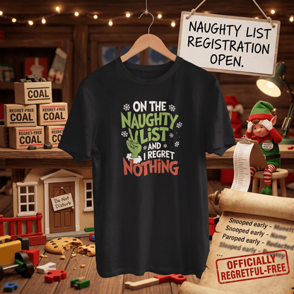 A navy graphic t-shirt hanging on a wooden hanger in a rustic holiday-themed room setting. The shirt features a festive graphic with the text 'ON THE NAUGHTY LIST AND I REGRET NOTHING' in white, green, and red lettering, accompanied by a Grinch face and snowflake details.