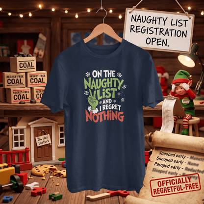 A navy blue graphic t-shirt hanging on a wooden hanger in a cozy, decorated workshop setting. The shirt features a festive graphic that reads 'ON THE NAUGHTY LIST AND I REGRET NOTHING' in white, green, and red lettering, accompanied by a green stocking and snowflake details.
