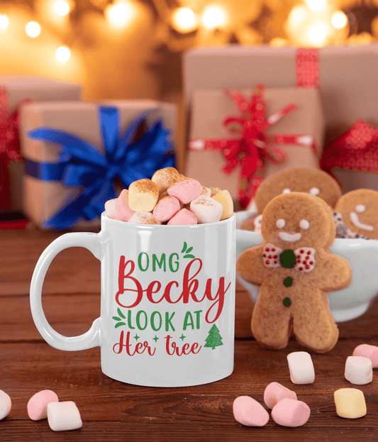 A white ceramic mug filled with marshmallows sits on a wooden table in a cozy holiday setting. The mug has a festive red and green printed message reading 'OMG Becky LOOK AT Her tree' with a small tree icon.
