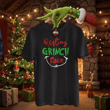 A black graphic t-shirt featuring the text 'Resting GRINCH Face' in red, green, and white. The shirt is shown hanging on a wooden hanger decorated with a green Grinch hand, Christmas lights, and a Santa hat, set against a cozy, warmly lit Christmas living room background with a tree, fireplace, and gifts.