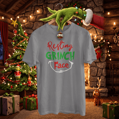 A light gray t-shirt featuring the text 'Resting GRINCH Face' in red and green lettering, hanging on a wooden hanger with a Grinch-themed Santa hat and ornament. The background depicts a cozy, festive Christmas living room with a decorated tree, fireplace, and gifts.