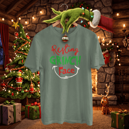 A sage green t-shirt featuring a 'Resting GRINCH Face' graphic, hanging on a wooden hanger decorated with a Christmas ornament and a Grinch hand wearing a Santa hat. The scene is set in a cozy, festive Christmas living room with a decorated tree, fireplace, and gifts.