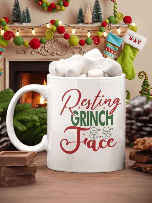 A white ceramic mug filled with marshmallows sits on a wooden table in a cozy holiday setting with a fireplace, garlands, stockings, and cookies. The mug is printed with the phrase 'Resting Grinch Face' in red and green lettering.