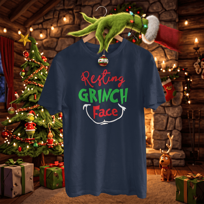 A navy blue graphic t-shirt featuring the text 'Resting GRINCH Face' in red, green, and white. The shirt is displayed on a hanger decorated with a Christmas ornament, surrounded by a festive Christmas tree, stockings, and a fireplace in the background.