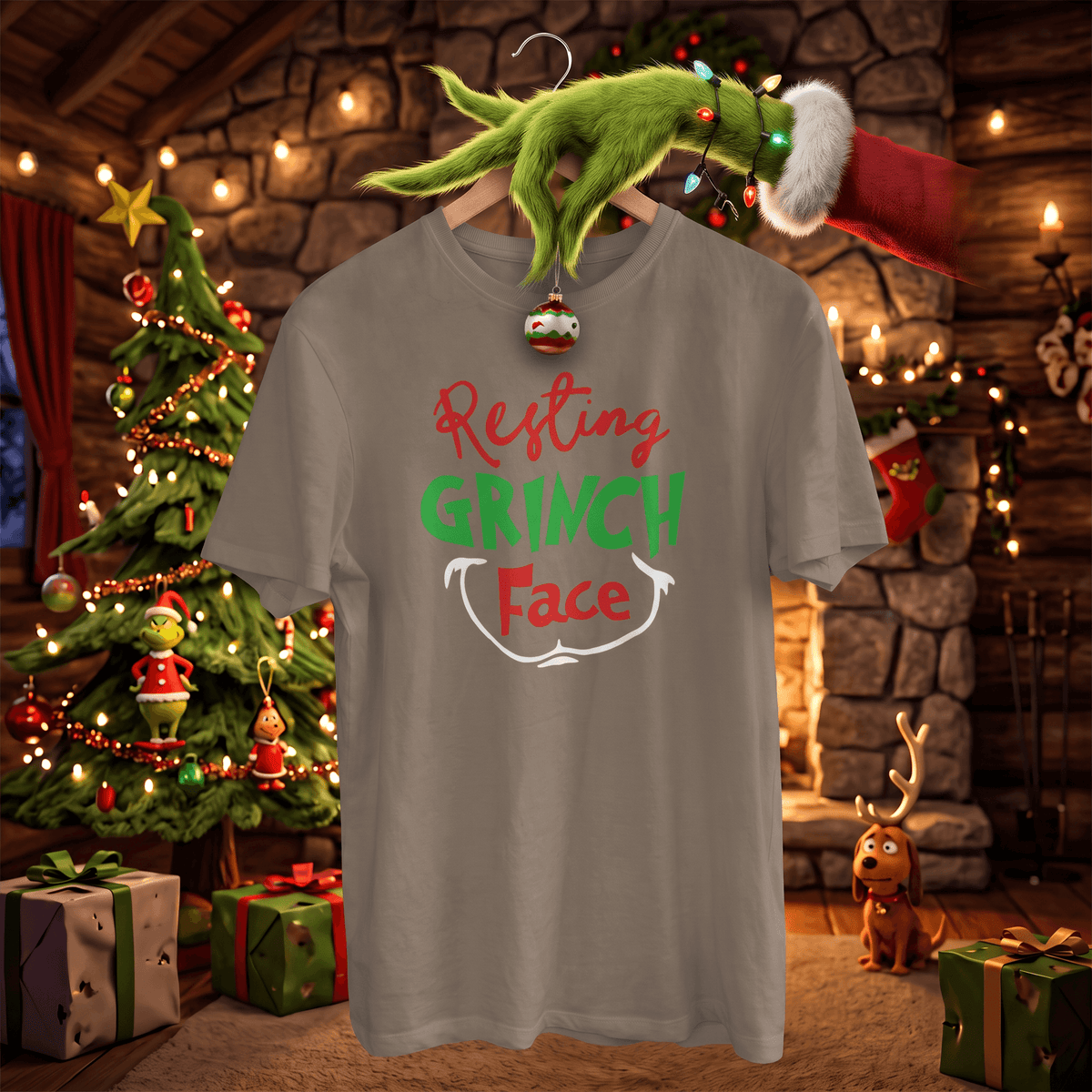A gray graphic t-shirt featuring the text 'Resting GRINCH Face' in red, green, and white, hanging on a wooden hanger with a Grinch-themed green hand and Santa hat decoration. The background is a cozy, festive Christmas cabin scene with a decorated tree, warm lights, and gifts.