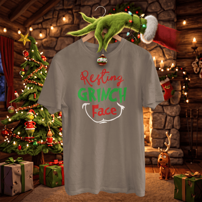 A gray graphic t-shirt featuring the text 'Resting GRINCH Face' in red, green, and white, hanging on a wooden hanger with a Grinch-themed green hand and Santa hat decoration. The background is a cozy, festive Christmas cabin scene with a decorated tree, warm lights, and gifts.