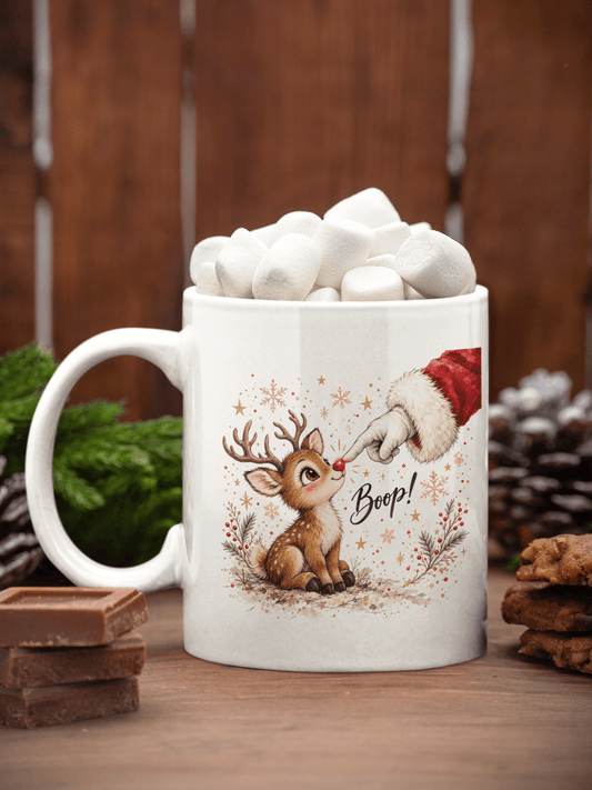 A white ceramic mug filled with marshmallows, featuring a festive illustration of a cute brown reindeer sitting next to Santa's hand with the word 'Boop!' written in script. The mug is set on a wooden surface with chocolate bars, pinecones, and greenery against a rustic wooden background.