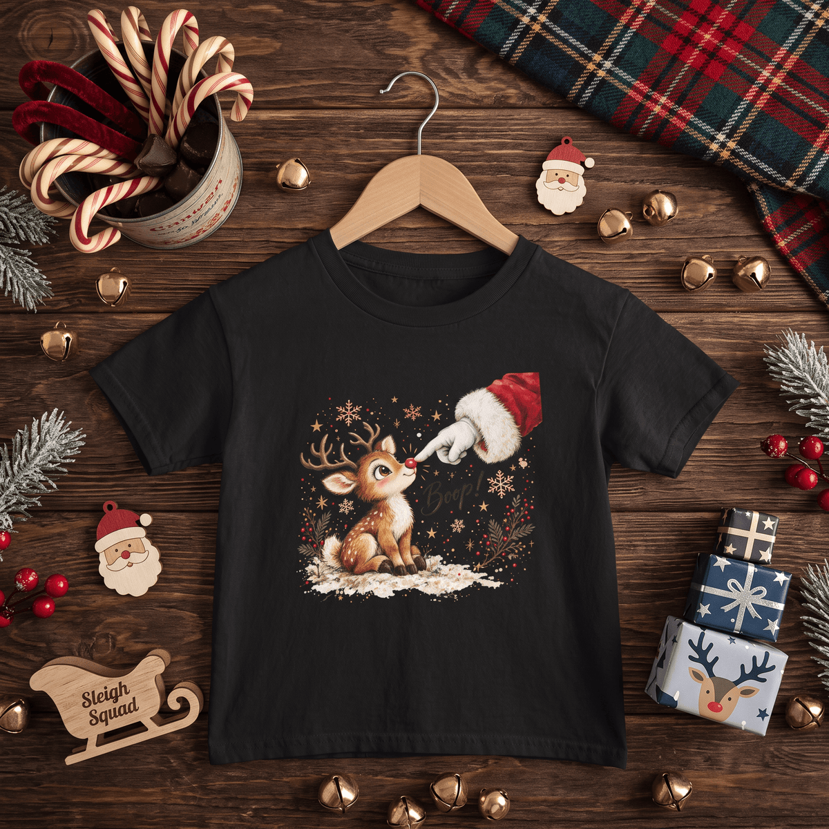 A black short-sleeve graphic t-shirt on a wooden hanger, styled in a Christmas-themed flat lay with candy canes, a plaid scarf, gold bells, pine branches, wrapped gifts, and a wooden sleigh on a dark wood background.