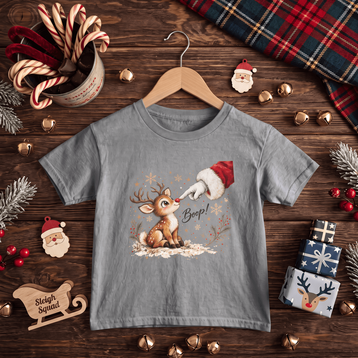 A light gray short-sleeve t-shirt on a wooden hanger, featuring a Christmas graphic of a reindeer and Santa's boot. The shirt is styled in a flat-lay arrangement on a dark wooden table with holiday decorations including candy canes, pine branches, gold bells, and wrapped gifts.