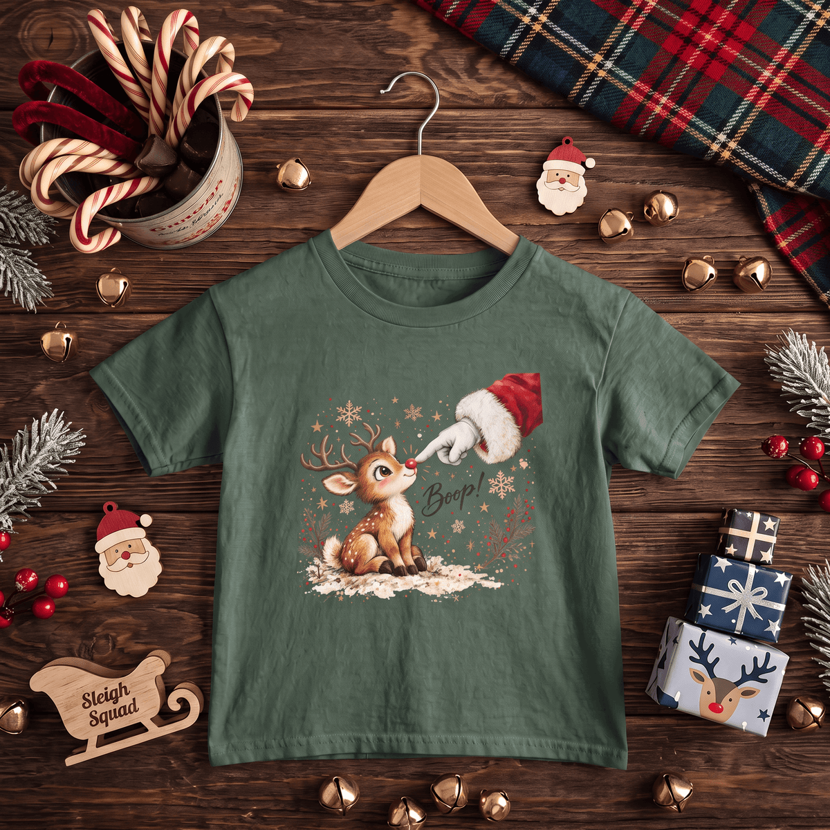A green short-sleeve kids' t-shirt with a Christmas graphic of a deer and Santa, displayed on a wooden hanger over a rustic Christmas-themed flat-lay background.