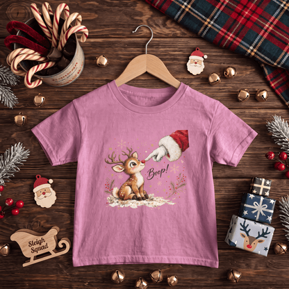 A pink short-sleeve t-shirt on a wooden hanger, featuring a Christmas graphic of a reindeer and Santa's boot. The shirt is displayed in a flat-lay arrangement on a dark wooden table surrounded by holiday decorations including candy canes, pine branches, gold bells, and a plaid scarf.