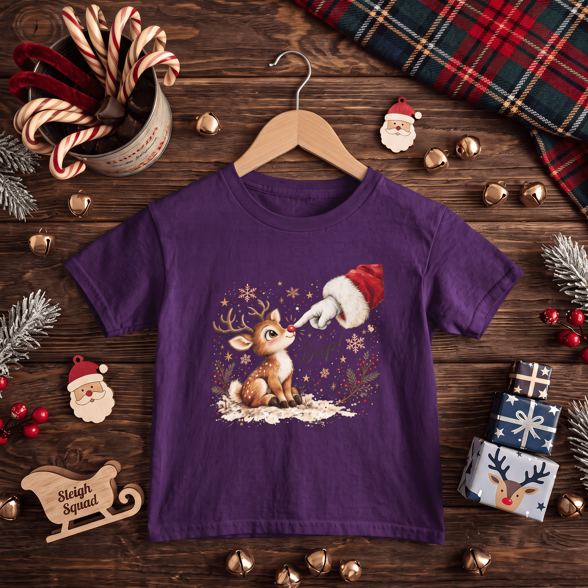 A purple short-sleeve kids' t-shirt with a Christmas graphic of a reindeer and Santa's boot, displayed on a wooden hanger over a rustic Christmas-themed flat-lay background.