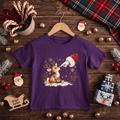A purple short-sleeve kids' t-shirt with a Christmas graphic of a reindeer and Santa's boot, displayed on a wooden hanger over a rustic Christmas-themed flat-lay background.