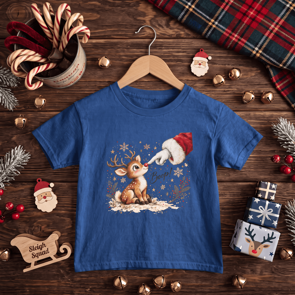 A blue short-sleeve kids' t-shirt on a wooden hanger, decorated with a festive Christmas graphic of a reindeer and Santa's hat. The shirt is styled in a flat-lay on a dark wooden table surrounded by holiday decorations including candy canes, pine branches, gold bells, and wrapped gifts.