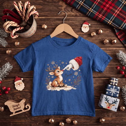 A blue short-sleeve kids' t-shirt on a wooden hanger, decorated with a festive Christmas graphic of a reindeer and Santa's hat. The shirt is styled in a flat-lay on a dark wooden table surrounded by holiday decorations including candy canes, pine branches, gold bells, and wrapped gifts.