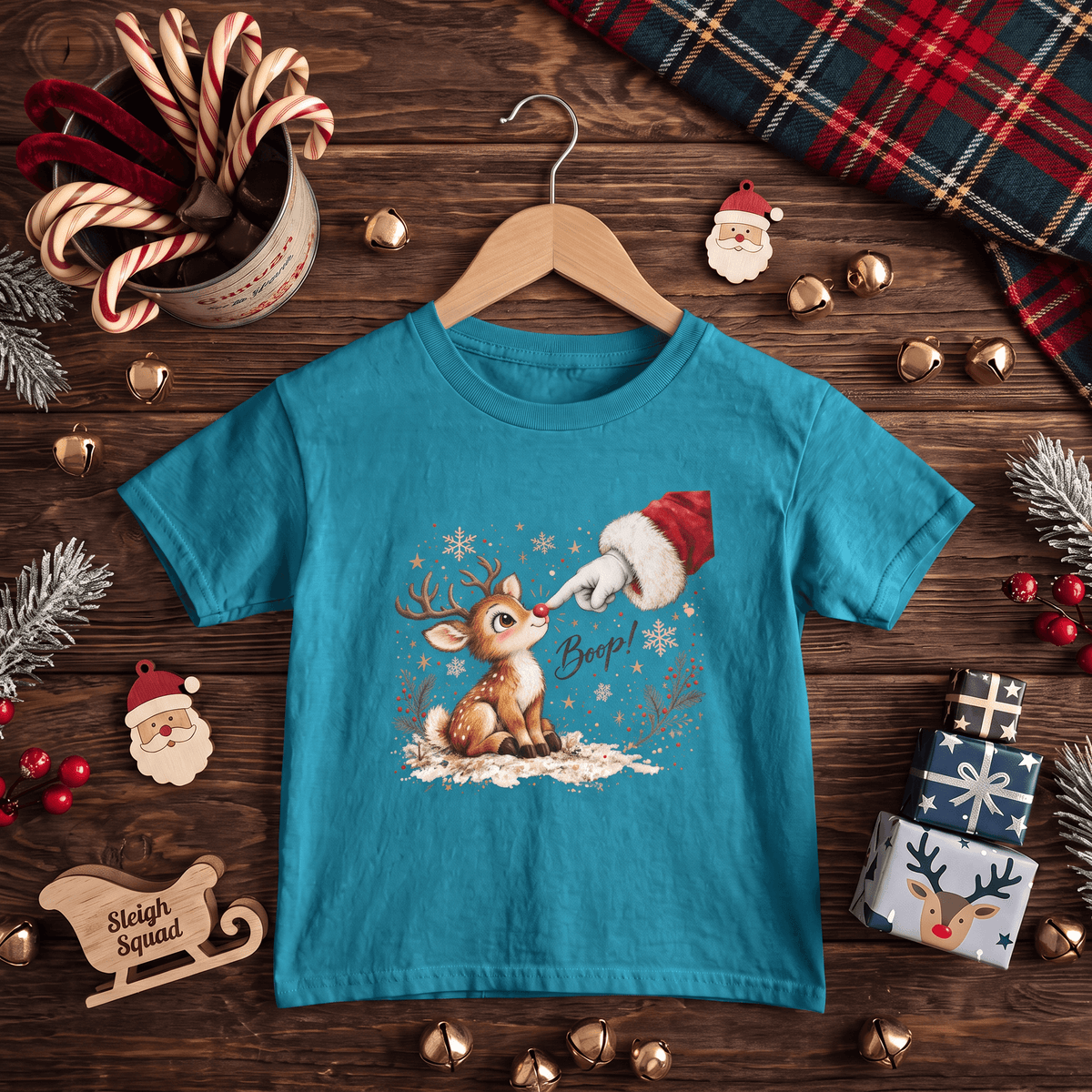 A turquoise short-sleeve t-shirt on a wooden hanger, decorated with a Christmas graphic of a reindeer and Santa. The shirt is styled in a flat-lay on a dark wooden background with holiday decorations including candy canes, pinecones, gold bells, a plaid scarf, and a wooden sleigh.