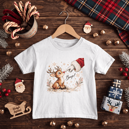 A white short-sleeve kids' T-shirt on a wooden hanger, featuring a Christmas graphic of a reindeer with antlers sniffing a Santa hat, with the word 'Boop!' written below. The shirt is displayed on a wooden surface surrounded by holiday props including candy canes, pine branches, gold bells, a plaid scarf, and small Santa decorations.