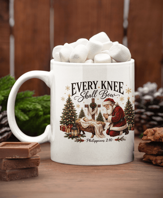 A white ceramic mug filled with marshmallows sits on a wooden surface. The mug features a Christmas-themed illustration with Santa kneeling by the baby Jesus, Christmas trees, gifts, and the text 'EVERY KNEE Shall Bow' and 'Philippians 2:10'.