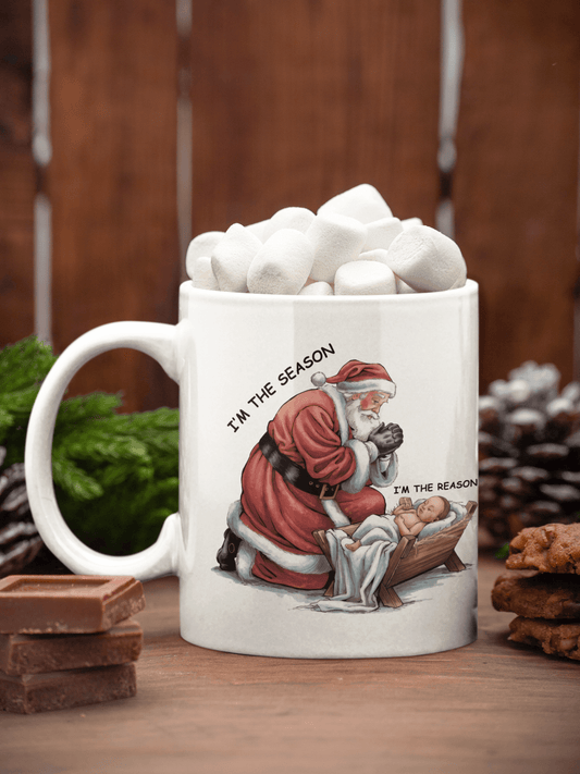 A white ceramic mug filled with marshmallows sits on a wooden surface. The mug features a printed illustration of Santa kneeling by a baby in a manger, with the text 'I'M THE SEASON' around the artwork.