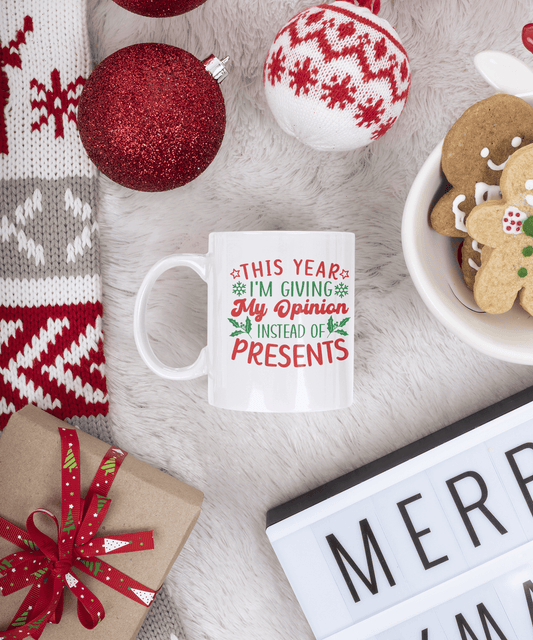 A white ceramic mug with a humorous Christmas message printed in red and green text, styled in a cozy holiday flat-lay with a knitted scarf, ornaments, gingerbread cookies, and a wrapped gift on a white faux fur surface.