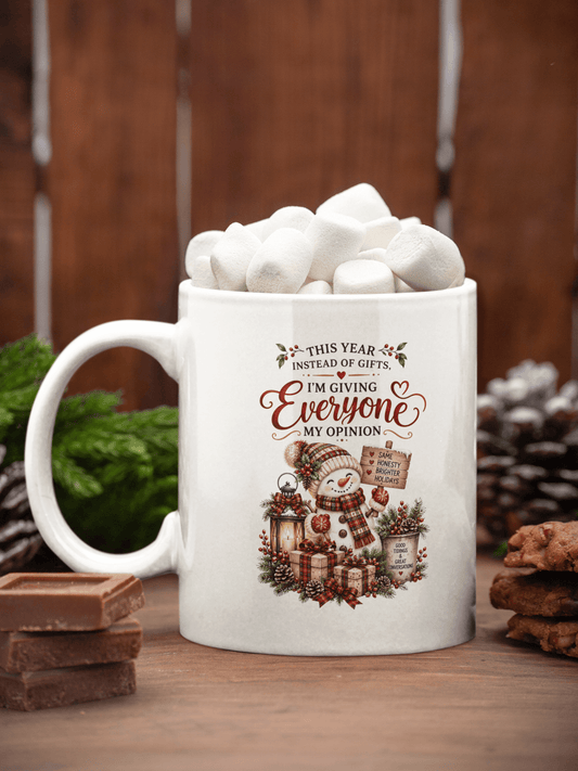A white ceramic mug filled with marshmallows and hot chocolate, placed on a wooden surface with chocolate bars, pinecones, and greenery in a rustic holiday setting. The mug features a printed design of a snowman and the text, 'THIS YEAR INSTEAD OF GIFTS I'M GIVING Everyone MY OPINION'.